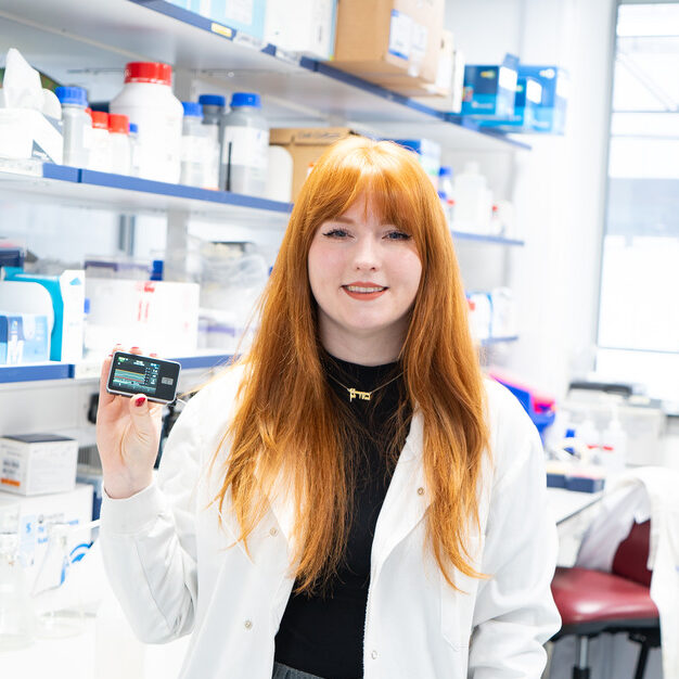 A photo of Morgan Shaw wearing a lab coat in a lab and holding up her insulin pump.