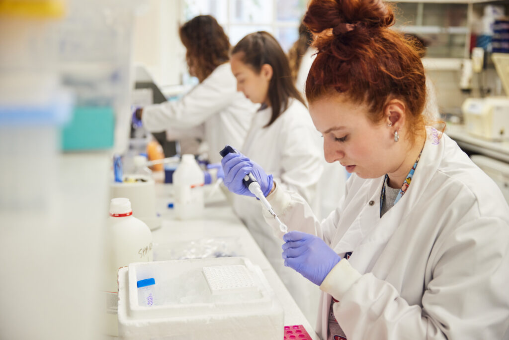Three female researchers working at a lab bench wearing white lab coats and purple gloves.